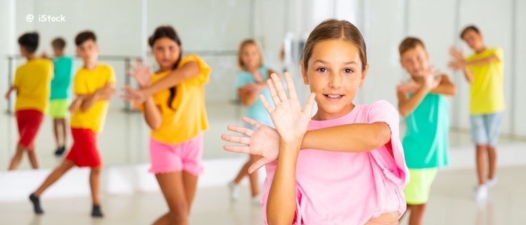 Eine Gruppe von Kindern in einem Tanzstudio, in farbenfroher Kleidung, übt Tanzschritte mit Lächeln im Gesicht.