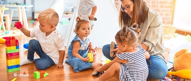 Eine Frau spielt mit einer Gruppe von Kindern in einem Spielzimmer. Die Kinder spielen mit Spielzeug auf dem Boden.