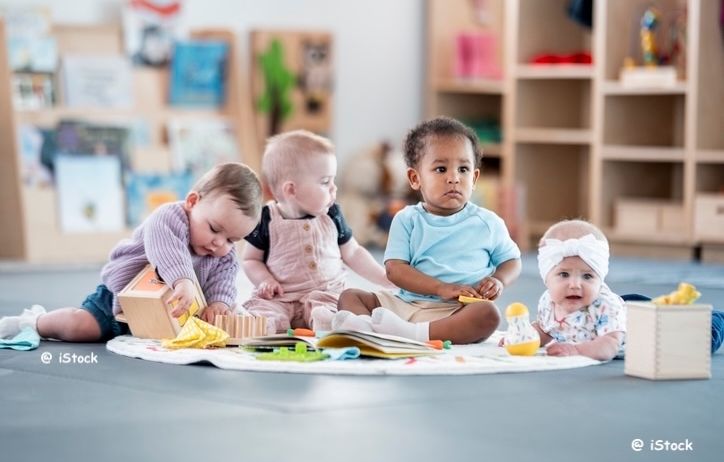 Fünf Babys sitzen auf dem Boden in einem Raum und spielen mit Spielzeug und Büchern. Der Raum hat Holzregale mit Büchern und Spielzeug. Der Boden ist mit einer weißen Matte bedeckt. Die Babys schauen sich gegenseitig an und scheinen sich zu amüsieren.