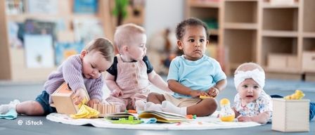 Fünf Babys sitzen auf dem Boden in einem Raum und spielen mit Spielzeug und Büchern. Der Raum hat Holzregale mit Büchern und Spielzeug. Der Boden ist mit einer weißen Matte bedeckt. Die Babys schauen sich gegenseitig an und scheinen sich zu amüsieren.