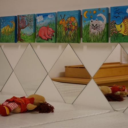 Eine Reihe bunter Gemälde hängt an einer weißen Wand. Unten liegt eine Puppe auf einer weißen Oberfläche, wobei ein Spiegel die Szene reflektiert.
