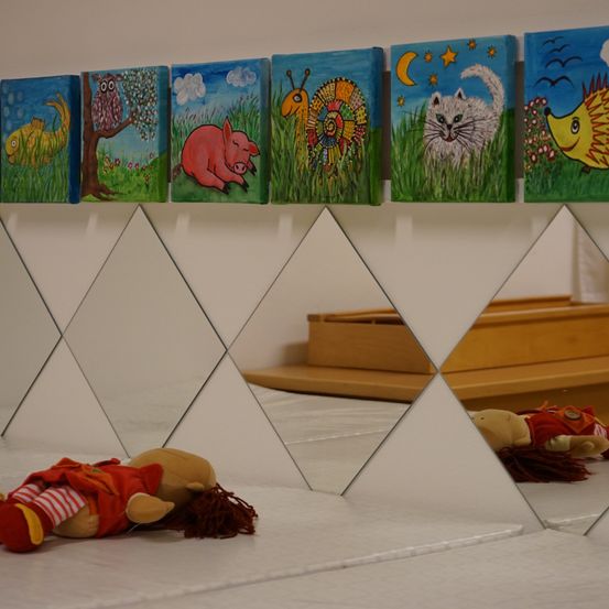 Eine Reihe bunter Gemälde hängt an einer weißen Wand. Unten liegt eine Puppe auf einer weißen Oberfläche, wobei ein Spiegel die Szene reflektiert.