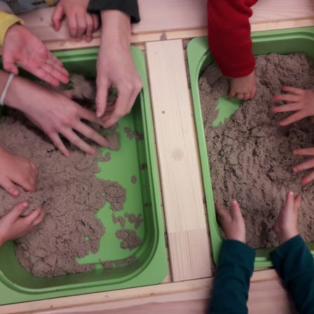 Eine Gruppe von Kindern spielt in Sandkästen. Die Kästen sind mit Sand gefüllt und auf einer Holzoberfläche platziert. Jedes Kind ist damit beschäftigt, mit den Händen im Sand zu spielen.