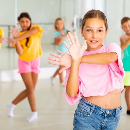 Eine Gruppe von Kindern in einem Tanzstudio, in farbenfroher Kleidung, übt Tanzbewegungen. Ein Mädchen in der Mitte lächelt in die Kamera.