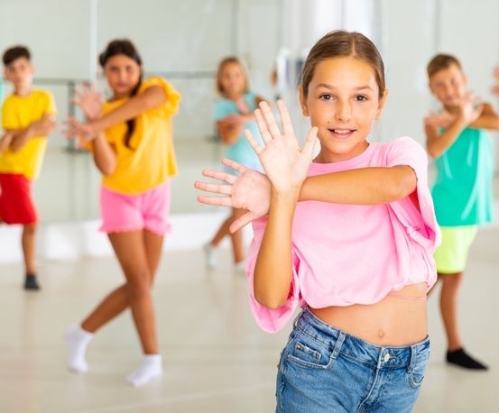 Eine Gruppe von Kindern in einem Tanzstudio, in farbenfroher Kleidung, übt Tanzbewegungen. Ein Mädchen in der Mitte lächelt in die Kamera.
