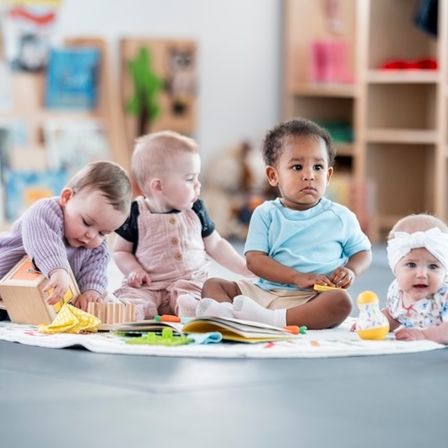 Fünf Babys sitzen auf dem Boden in einem Raum und spielen mit Spielzeug und Büchern. Sie sind von Regalen umgeben, die mit Büchern und Spielzeug gefüllt sind.