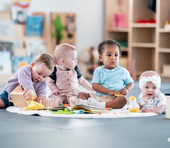 Fünf Babys sitzen auf dem Boden in einem Raum und spielen mit Spielzeug und Büchern. Sie sind von Regalen umgeben, die mit Büchern und Spielzeug gefüllt sind.