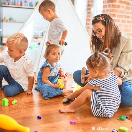 Eine Frau und mehrere Kinder spielen mit Spielzeug auf einem Holzboden. Die Kinder sitzen auf dem Boden und die Frau kniet. Die Frau lächelt und schaut die Kinder an. Die Kinder spielen mit Spielzeug und einige von ihnen stehen. Der Raum hat eine Ziegelwand und ein weißes Zelt.