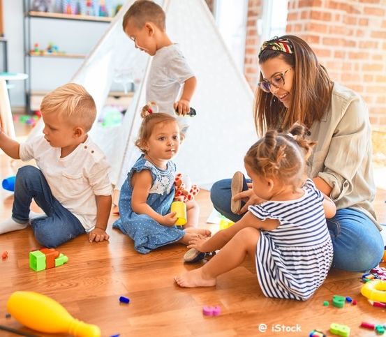 Eine Frau und mehrere Kinder spielen mit Spielzeug auf einem Holzboden. Die Kinder sitzen auf dem Boden und die Frau kniet. Die Frau lächelt und schaut die Kinder an. Die Kinder spielen mit Spielzeug und einige von ihnen stehen. Der Raum hat eine Ziegelwand und ein weißes Zelt.