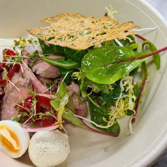 Ein weißer Teller mit einem Salat aus Blattgemüse, geschnittenem Fleisch und einem halbgekochten Ei. Das Gericht ist mit einem knusprigen Crouton garniert.