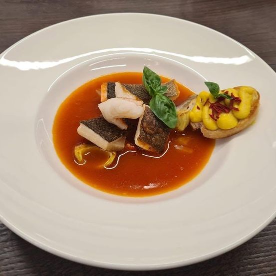 Ein weißer Teller mit einem Gericht aus Fischfilets in roter Sauce, garniert mit Basilikumblättern und einer gelben Aufstrich mit roten Toppings.