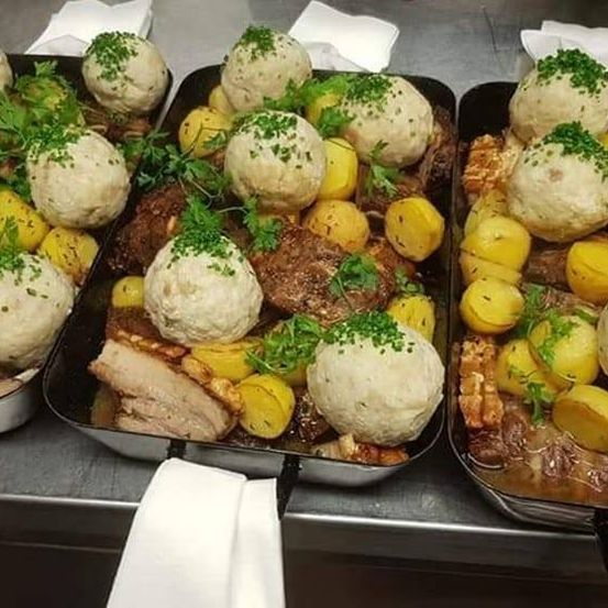 Drei Tabletts mit Essen, einschließlich Fleisch, Kartoffeln und Knödeln, mit grünen Kräutern garniert, werden auf einer Metalloberfläche präsentiert.