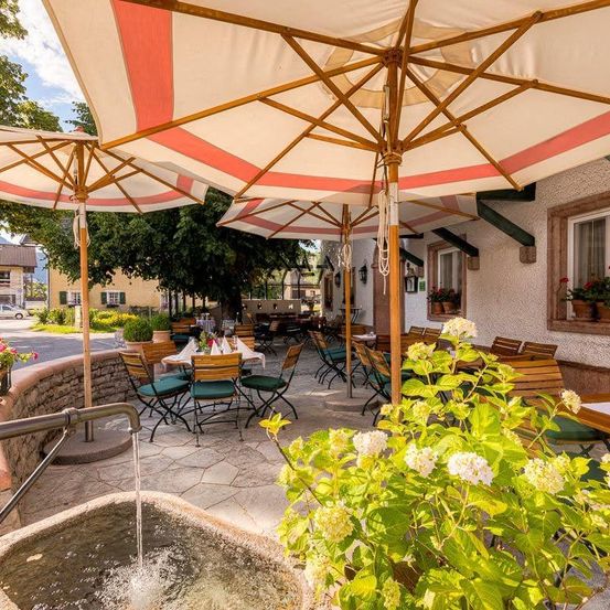 Ein Restaurant mit Außenbestuhlung unter großen Sonnenschirmen, umgeben von Grün und einem Brunnen. Bäume und Gebäude im Hintergrund.