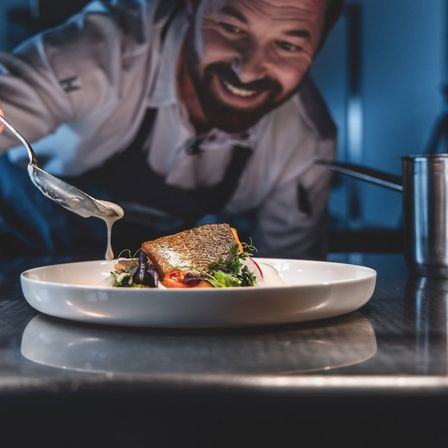 Ein Koch serviert ein Gericht mit Sauce, lächelt und schaut in die Kamera. Das Gericht ist auf einem weißen Teller mit Gemüse und einer Scheibe Brot.