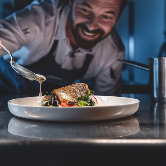 Ein Koch serviert ein Gericht mit Sauce, lächelt und schaut in die Kamera. Das Gericht ist auf einem weißen Teller mit Gemüse und einer Scheibe Brot.