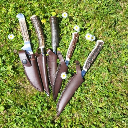 Fünf Messer mit braunen Lederscheiden liegen auf einem Grasfeld mit weißen Gänseblümchen verstreut.