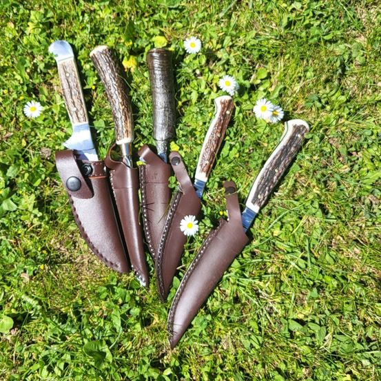 Fünf Messer mit braunen Lederscheiden liegen auf einem Grasfeld mit weißen Gänseblümchen verstreut.