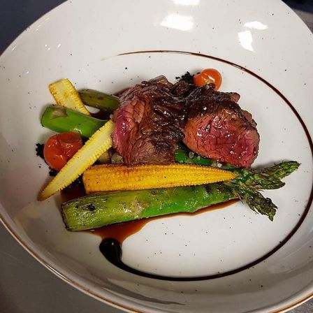 Ein weißer Teller enthält zwei Steaks mit Spargel, grünen Bohnen, Mais und Kirschtomaten. Der Teller hat einen braunen Rand und ein braunes Design am Rand.