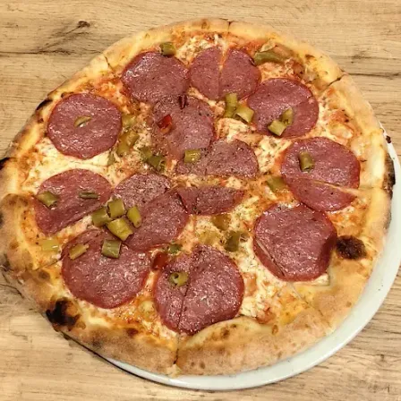 Eine ganze Pepperoni-Pizza mit grünen Pepperoni-Scheiben liegt auf einem weißen Teller, auf einem Holztisch.