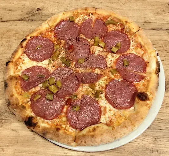 Eine ganze Pepperoni-Pizza mit grünen Pepperoni-Scheiben liegt auf einem weißen Teller, auf einem Holztisch.