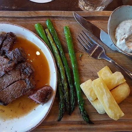 Ein Holztablett hält einen Teller mit geschnittenem Rindfleisch, einer Soße, Spargel und einer Schüssel mit Sahne. Ein Messer, eine Gabel und ein Glas befinden sich ebenfalls auf dem Tablett.