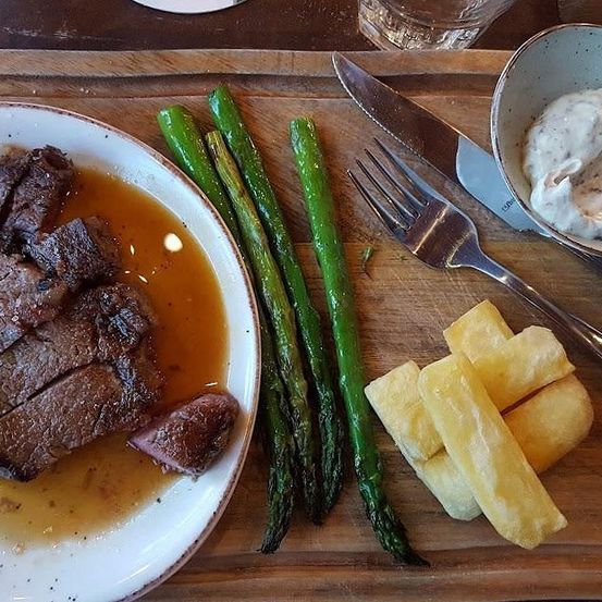Ein Holztablett hält einen Teller mit geschnittenem Rindfleisch, einer Soße, Spargel und einer Schüssel mit Sahne. Ein Messer, eine Gabel und ein Glas befinden sich ebenfalls auf dem Tablett.
