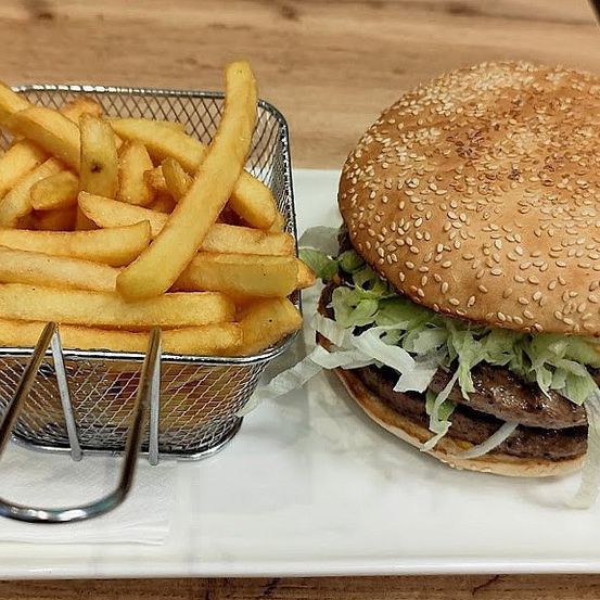 Ein Burger mit Salat und Sesamkörnern auf einem weißen Teller neben einem Korb Pommes Frites.