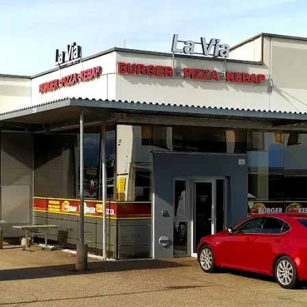 Ein rotes Auto steht vor einem La Via Restaurant mit einem Angebot an Burger, Pizza und Kebab. Das Gebäude ist weiß mit einem roten Vordach.