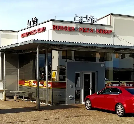 Ein rotes Auto steht vor einem La Via Restaurant mit einem Angebot an Burger, Pizza und Kebab. Das Gebäude ist weiß mit einem roten Vordach.