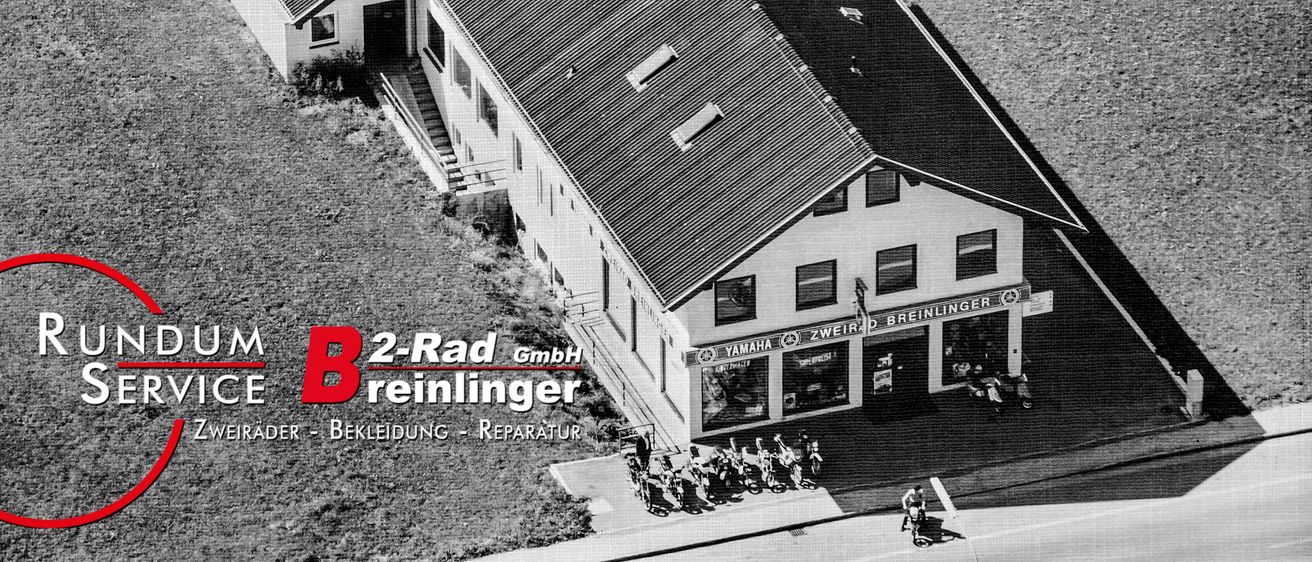 Ein Luftbild von einem Gebäude mit mehreren Motorrädern davor. Das Schild sagt 'Zweirad Breinlinger GmbH'. Das Gebäude hat ein geneigtes Dach und mehrere Fenster. Vor dem Gebäude stehen Leute.