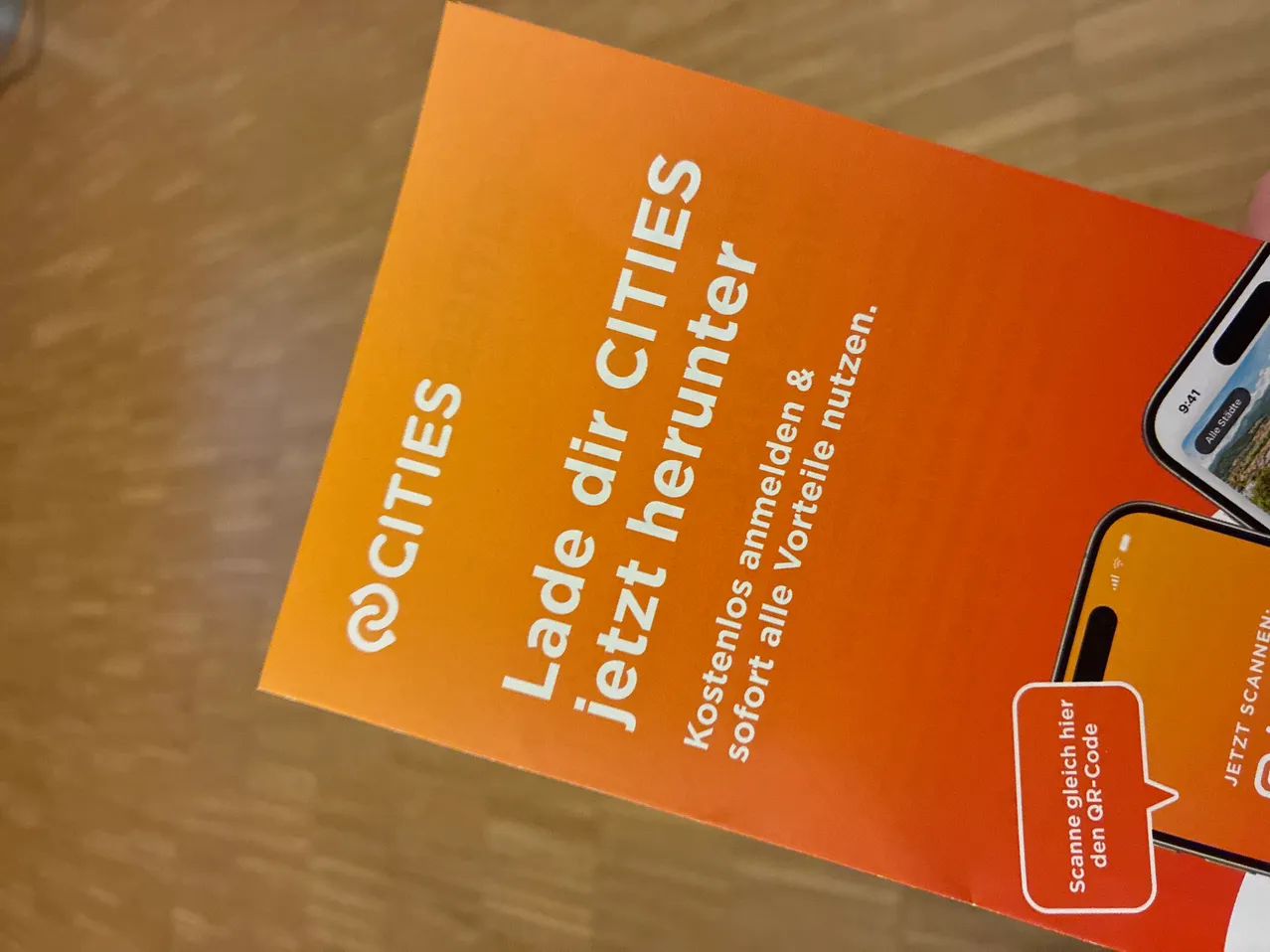 Ein Ausschnitt eines orangefarbenen Papiers mit weißem Text lautet '@CITIES Lade dir CITIES jetzt herunter'. Es bewirbt einen Stadtführer mit sofortigem Zugriff.