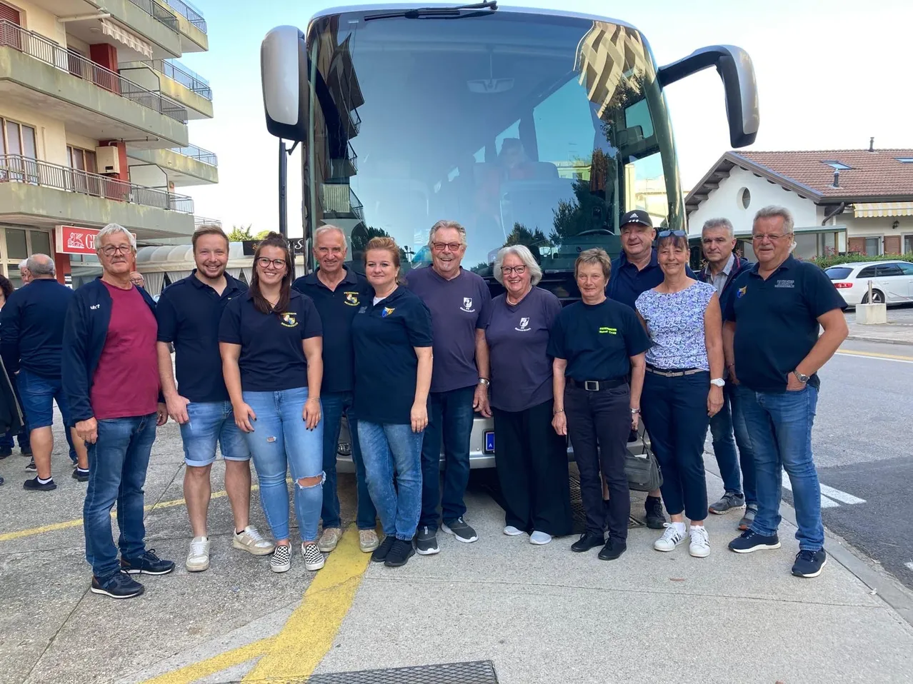 Eine Gruppe von Menschen in gleichen Shirts steht vor einem großen Bus. Die Umgebung scheint ein Parkplatz zu sein, mit Gebäuden im Hintergrund.