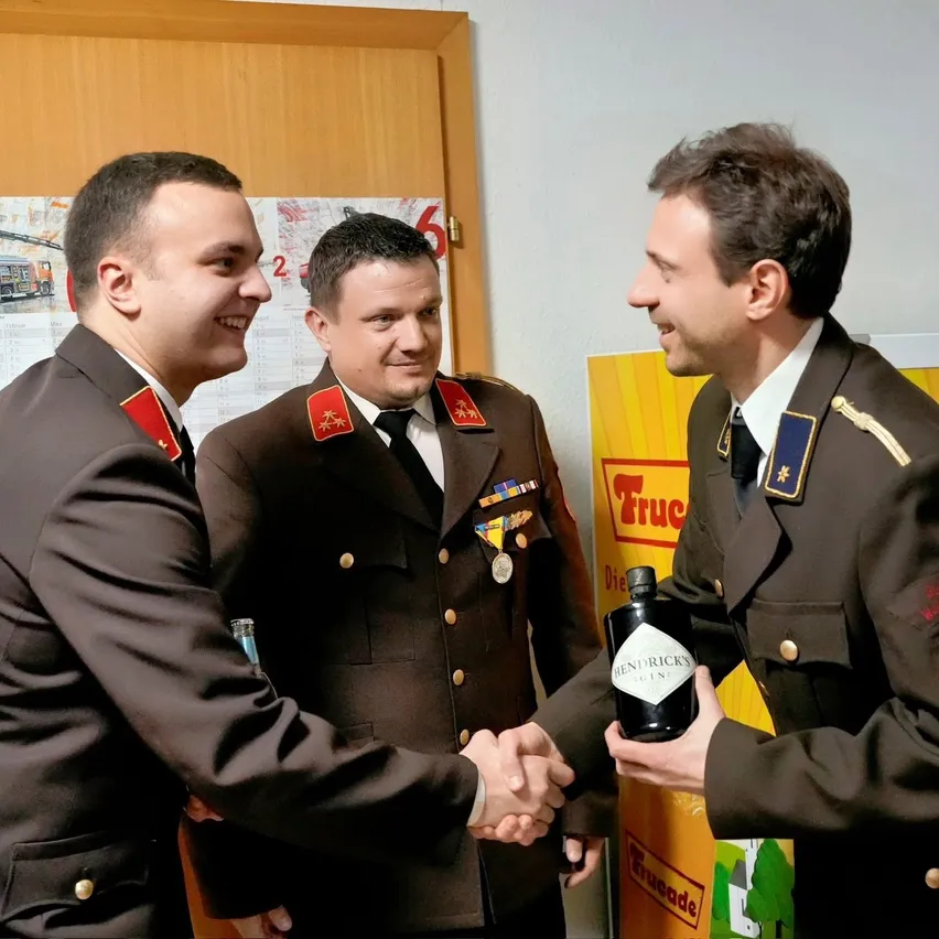 Drei Männer in Militäruniformen stehen und schütteln Hände. Der Mann auf der rechten Seite hält eine Flasche Hendrick's Gin.