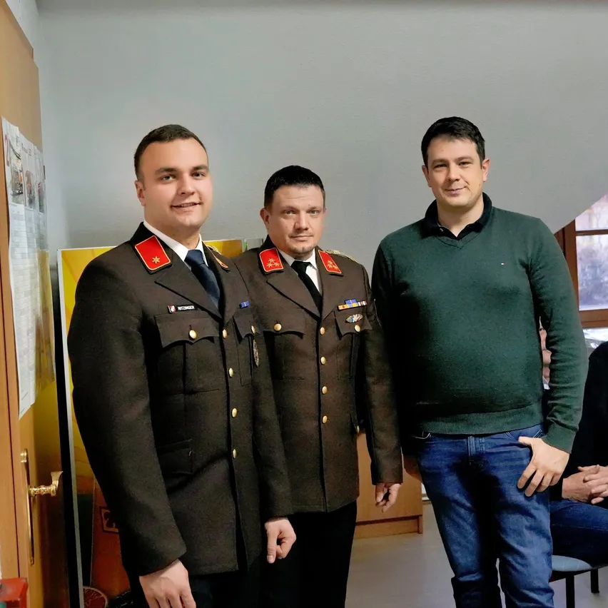Drei Männer in einem Raum, zwei in Militäruniformen und einer in einem grünen Pullover, stehen nebeneinander mit Lächeln auf den Gesichtern.