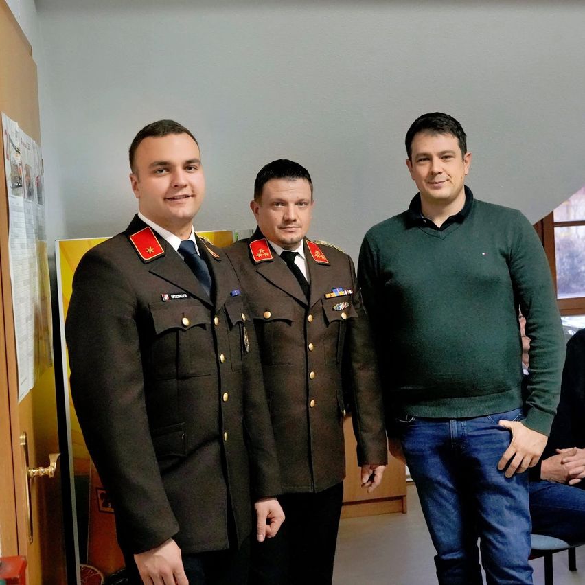 Three men in a room, two in military uniforms and one in a green sweater, standing next to each other with smiles on their faces.