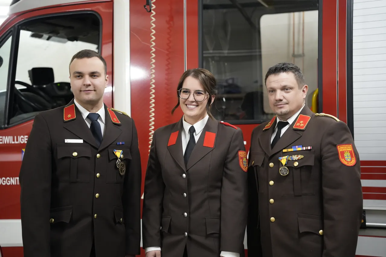 Drei Feuerwehrleute in Uniform stehen vor einem Feuerwehrwagen. Sie lächeln und haben Medaillen an ihren Uniformen.
