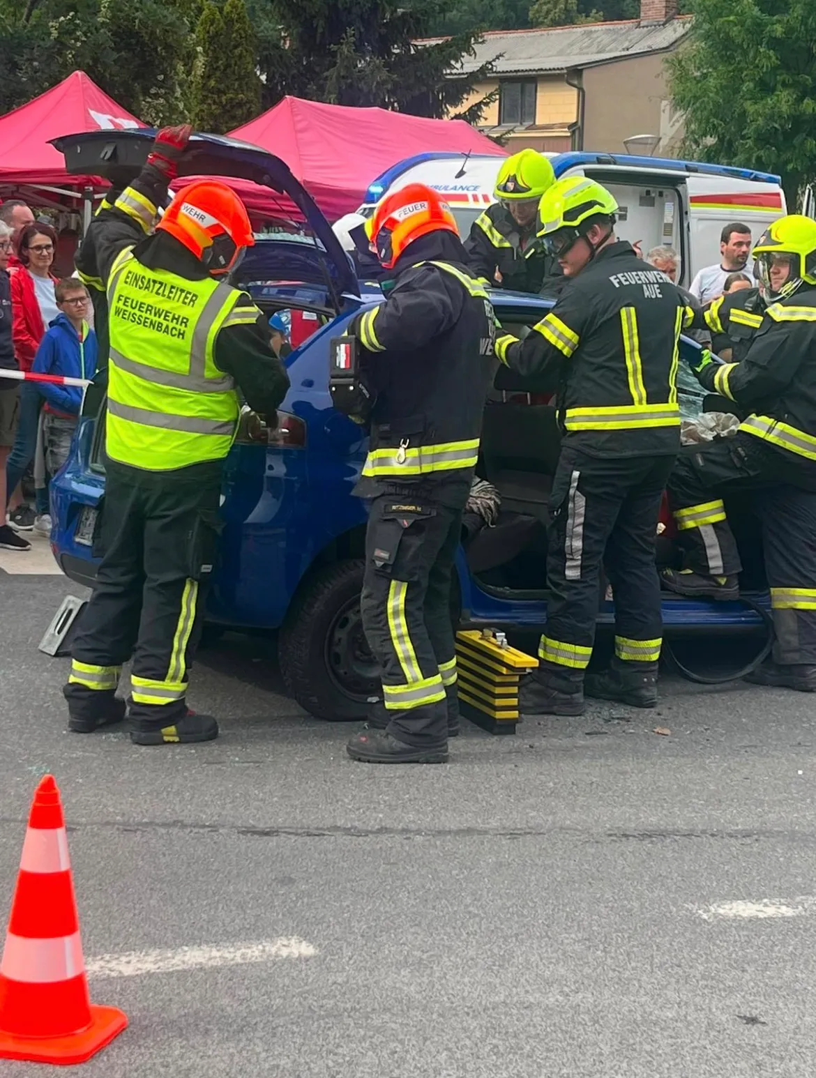 Mehrere Feuerwehrleute arbeiten an einem beschädigten Auto, tragen gelbe Westen und Helme. Sie sind draußen, mit Zuschauern im Hintergrund.