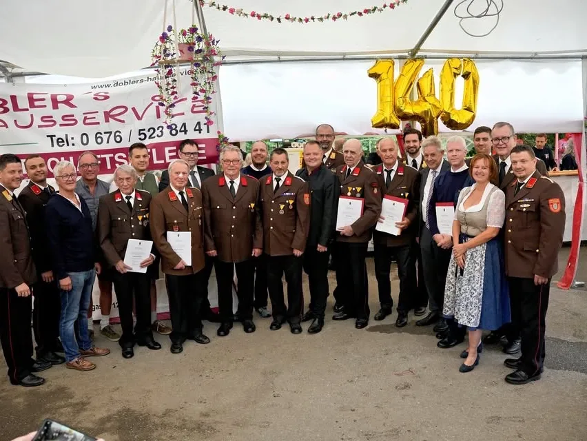 Gruppenfoto mit mehreren Männern und Frauen in Uniform, die Urkunden halten. Hinter ihnen steht ein weißes Zelt, das ein Banner und eine Ballonzahl 140 zeigt.