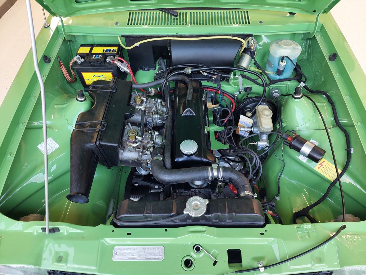 The interior of a green car displaying its engine and various components such as a black fuel tank, a blue coolant reservoir, and an array of wires and pipes.