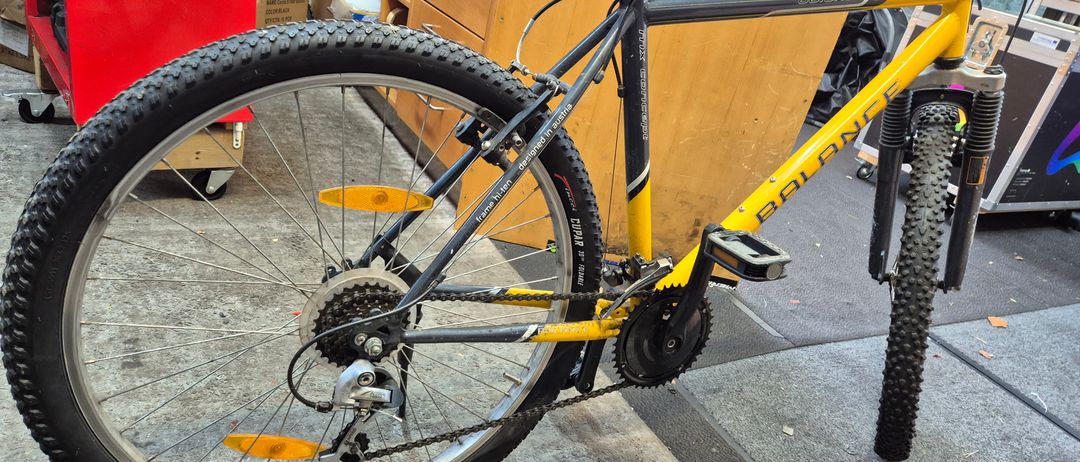 Ein gelbes Balance-Fahrrad steht in einer Werkstatt. Es hat einen schwarzen Rahmen, gelbe Reifen und eine Rückleuchte. Das Fahrrad steht auf einem Betonboden.