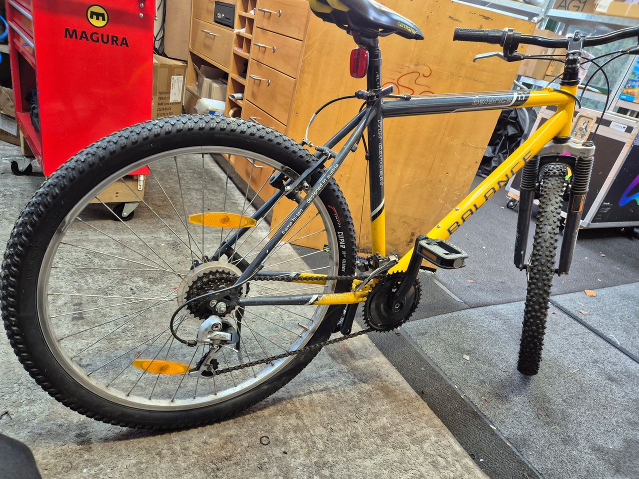 Ein gelbes BALANCE Mountainbike steht in einer Garage. Das Fahrrad hat schwarze und gelbe Reifen, einen schwarzen Sattel und ein rotes Licht hinten. Das Fahrrad steht auf einem grauen Boden. Hinter dem Fahrrad befindet sich ein hölzerner Schrank mit mehreren Schubladen und einer roten Aufbewahrungsbox.