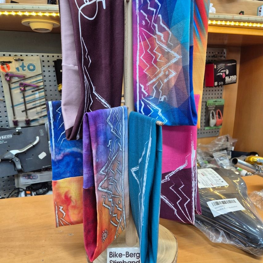 Ein hölzener Display mit verschiedenen farbenfrohen Bandanas darauf. Jedes Bandana hat einzigartige abstrakte Designs. Dahinter stehen Regale mit verschiedenen Artikeln, und der Boden ist mit mehr Gegenständen übersät.