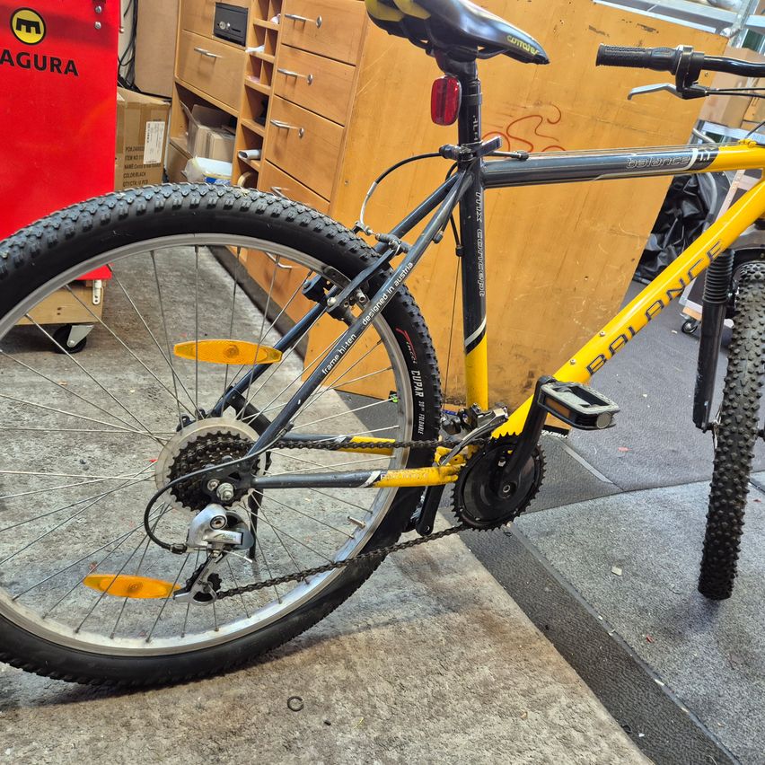 Ein gelbes und schwarzes Balance-Fahrrad steht in einer Garage. Es hat einen roten Reflektor und einen schwarzen Sattel. Im Hintergrund befinden sich Holzschränke und eine rote Lagerbox.