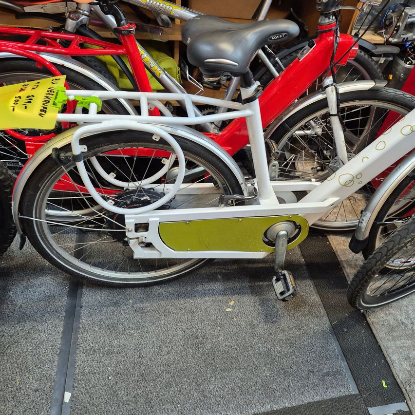 Ein weißes Fahrrad mit einem gelben Schild steht in einer Garage zwischen mehreren Fahrrädern. Es hat einen schwarzen Sattel und grüne Pedale.