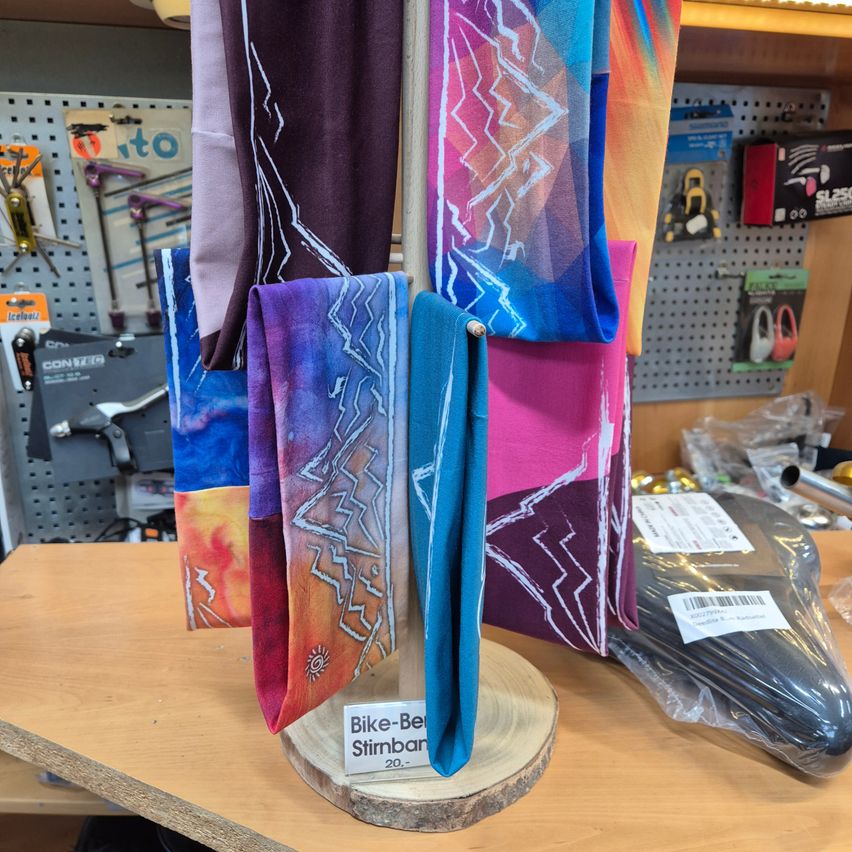 Bike-Be Stirnband 20 wird in einem Geschäft ausgestellt. Der Laden hat einen Holztisch mit verschiedenfarbigen Artikeln darauf. Hinter dem Tisch hängen mehrere Gegenstände an der Wand.