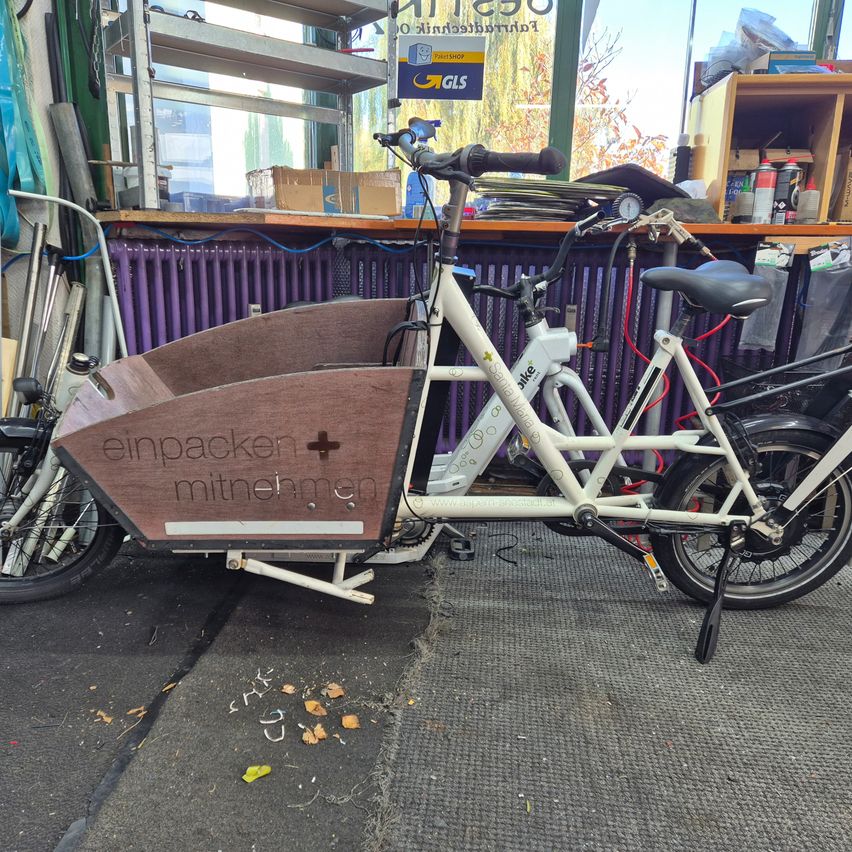 Ein weißer Lastenfahrrad mit braunen Kisten ist in einem Laden geparkt. Das Wort 'einpacker' steht auf der Kiste.