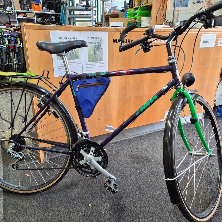 Ein lila und grünes Fahrrad steht in einem Geschäft mit einem hölzernen Tresen dahinter. Es hat eine blaue Tasche, die am Rahmen befestigt ist.