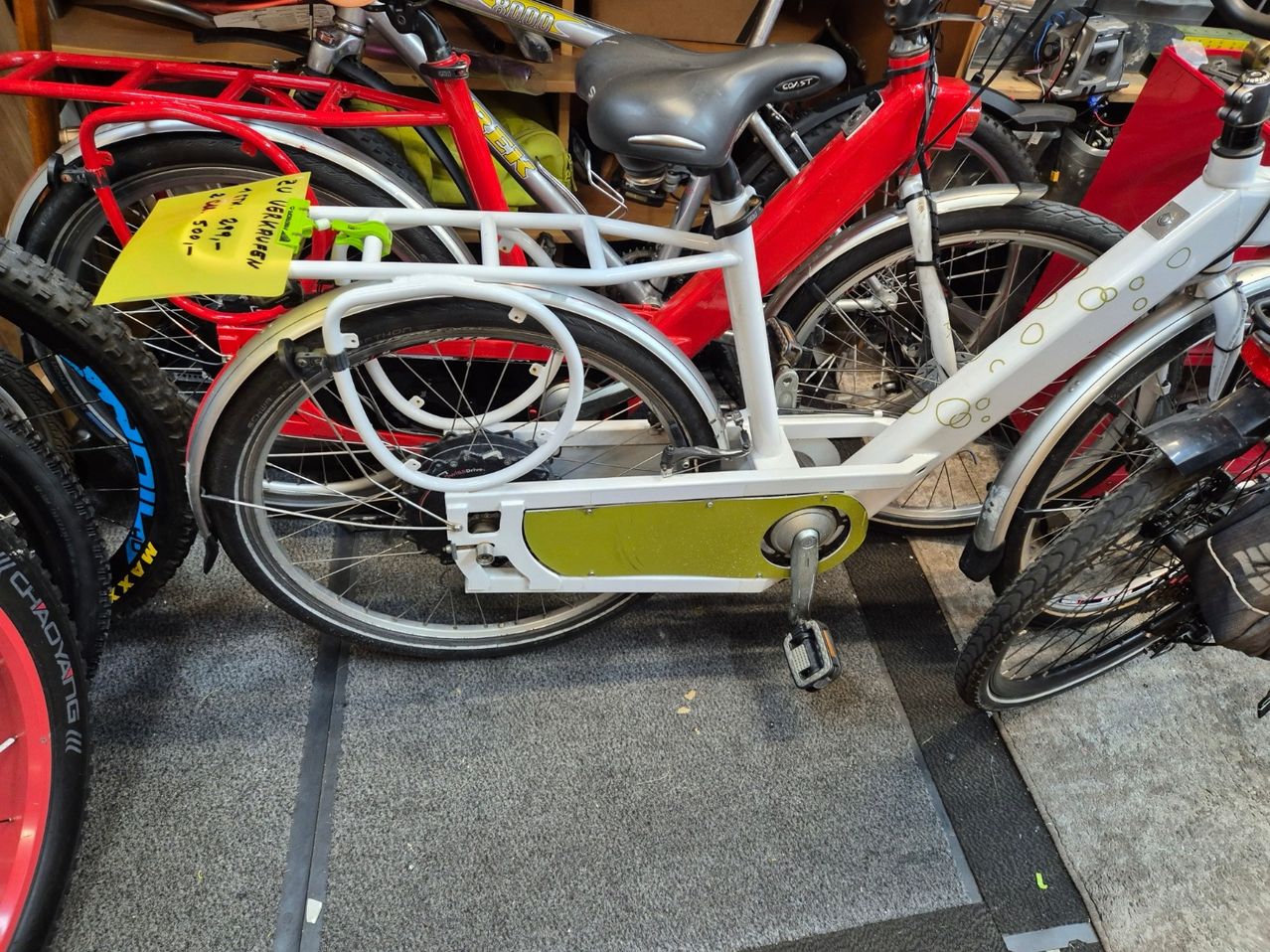 Ein weißes Fahrrad mit grünem Akku ist zwischen anderen Fahrrädern geparkt. Das Vorderrad hat einen gelben Anhänger.
