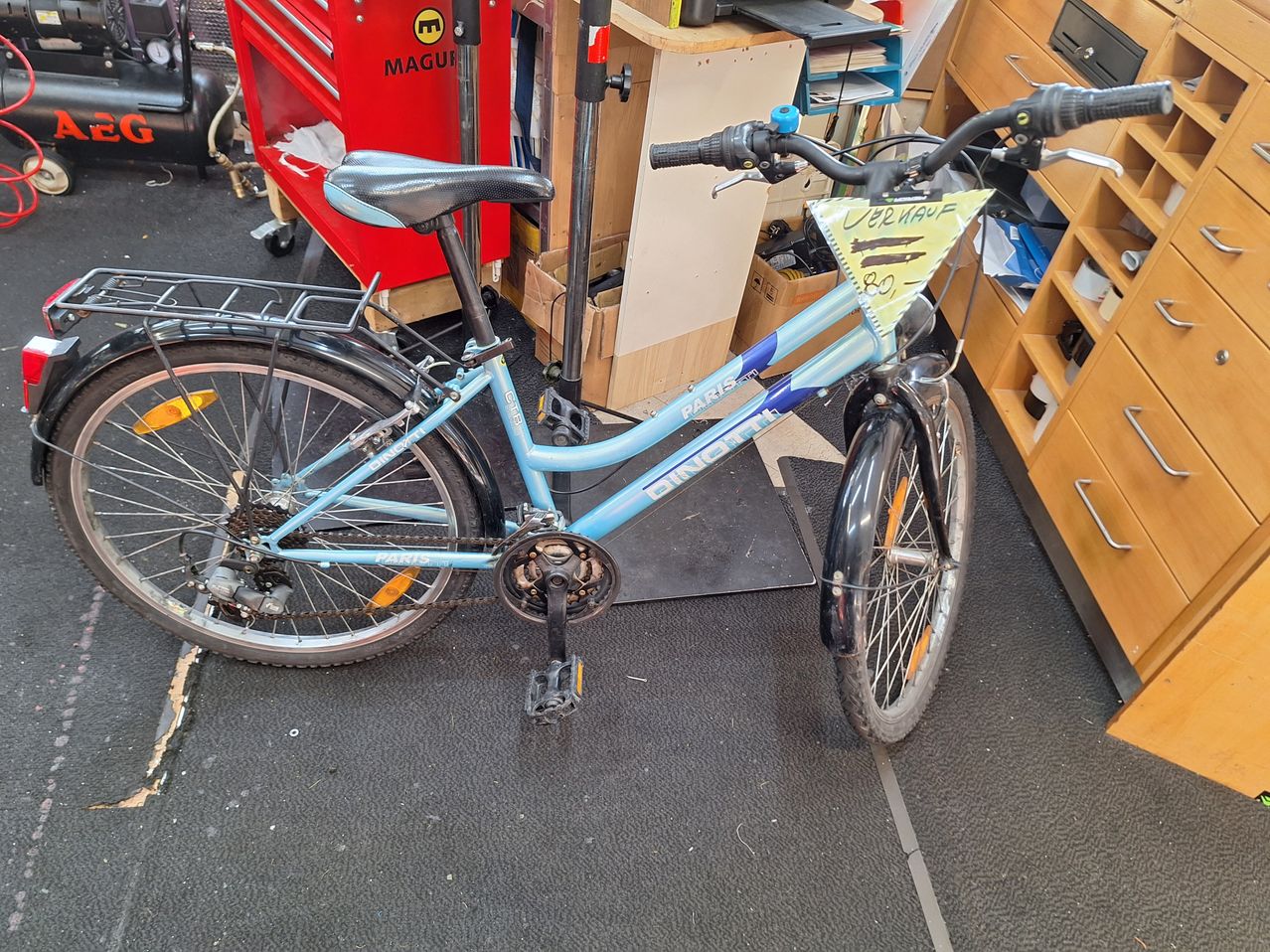 Ein blaues Fahrrad mit einem gelben Schild, auf dem 'Verkauf' steht, steht auf einem Teppichboden. Im Hintergrund befinden sich ein roter Werkzeugkasten und Holzregale.