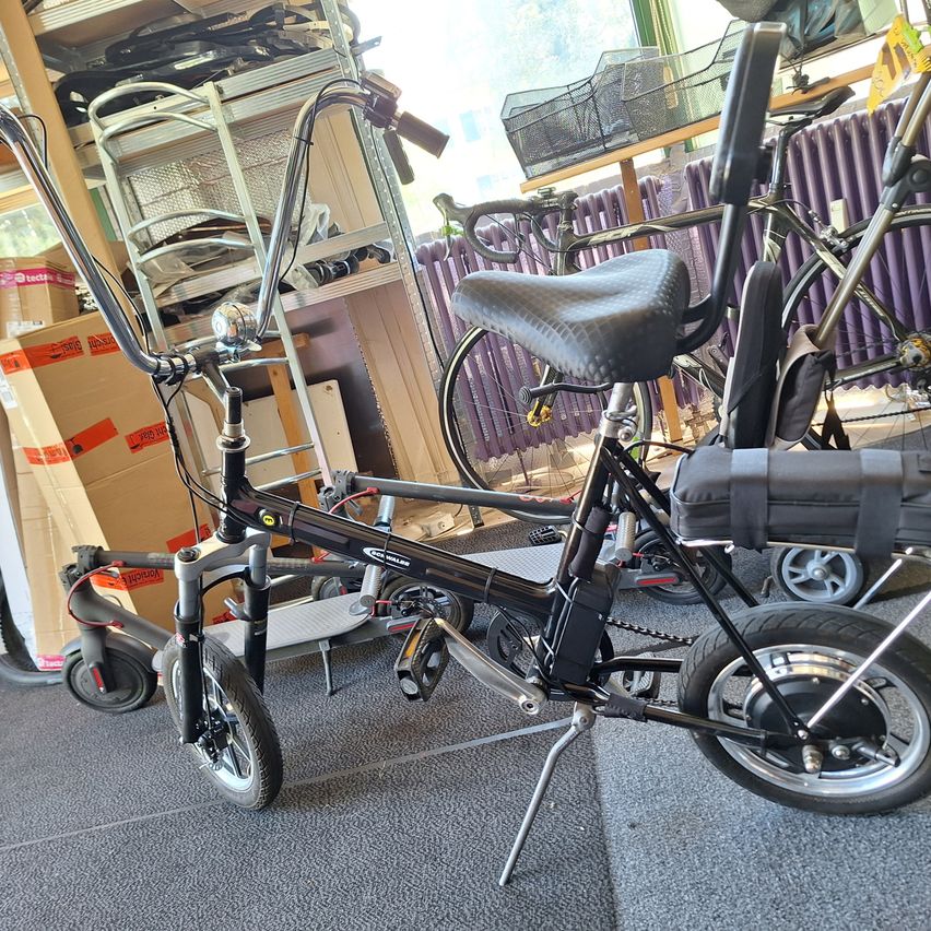 Ein schwarzes Fahrrad mit schwarzem Sattel und Ständer steht in einem Geschäft. Dahinter befinden sich weitere Fahrräder und Gegenstände.